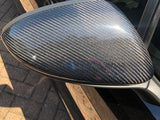 A car side mirror with a carbon fiber cover reflects light, attached to a black vehicle parked on a paved driveway, with sunlight creating a gloss effect.