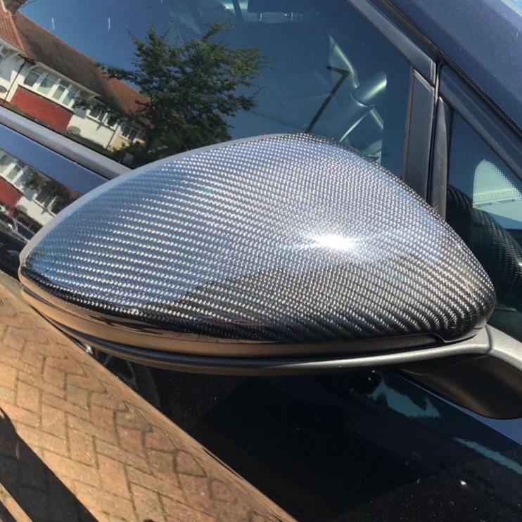 A car side mirror with a carbon fiber pattern is attached to a black vehicle. It reflects nearby houses and a tree on a sunny day, showing a suburban street scene.