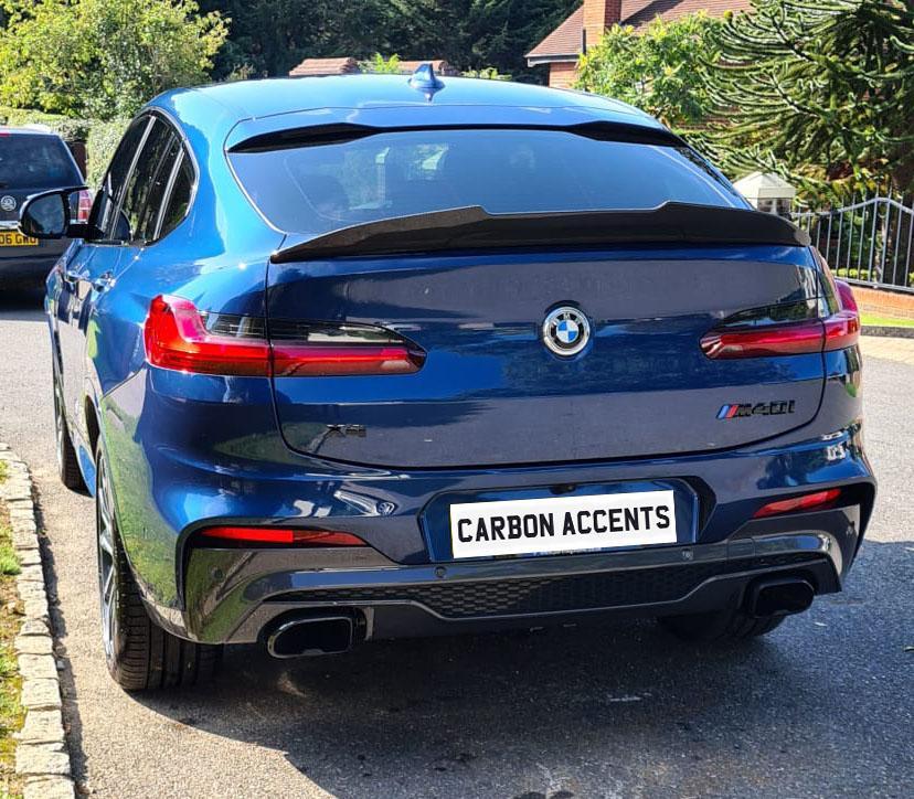 BMW car parked on a residential street with greenery visible. The license plate reads "CARBON ACCENTS." Nearby, there's another vehicle and houses in the background.
