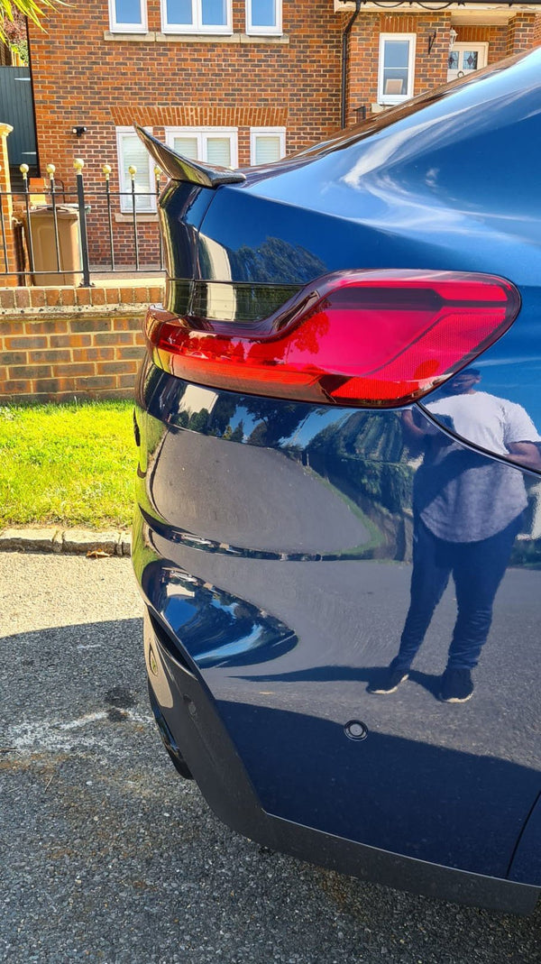 A blue car's rear with a red taillight is parked on a street. A person's reflection is visible on the car. A brick house and grass are in the background.