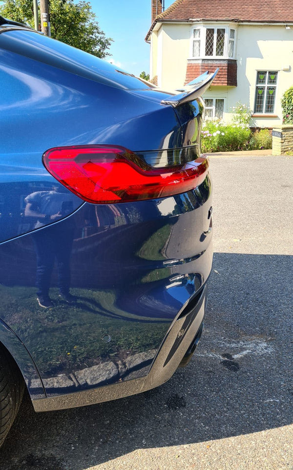 A blue car with a visible taillight and spoiler is parked on a sunny street, reflecting a residential house and greenery in its shiny surface.